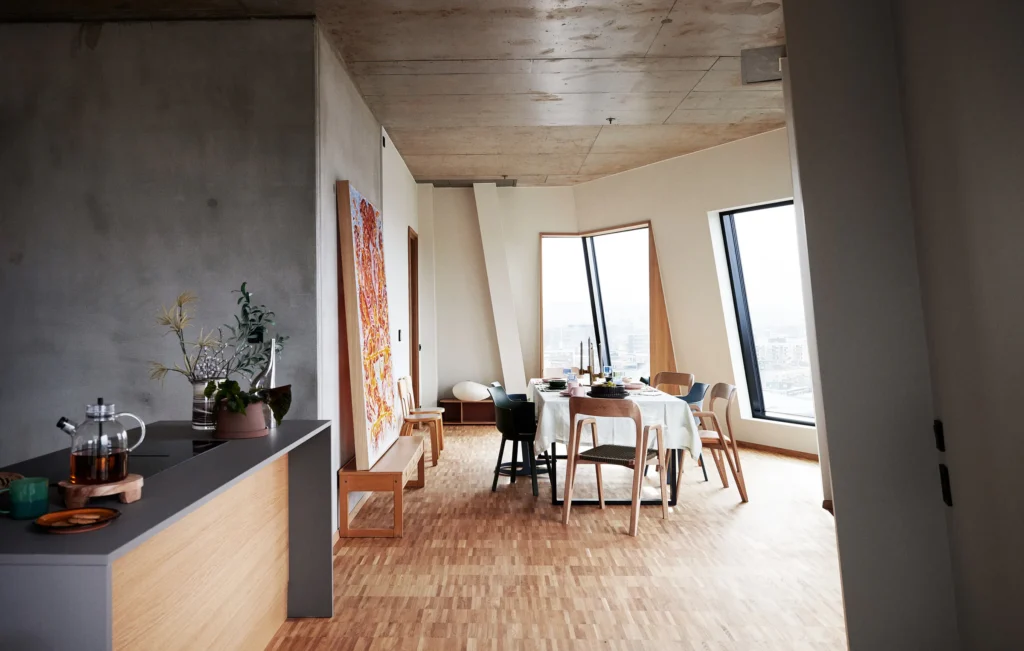 Modern Kitchen and dining area of one of the top floor apartments, with a wooden floor, a concrete ceiling, and two panoramic windows embedded in the angled facade
