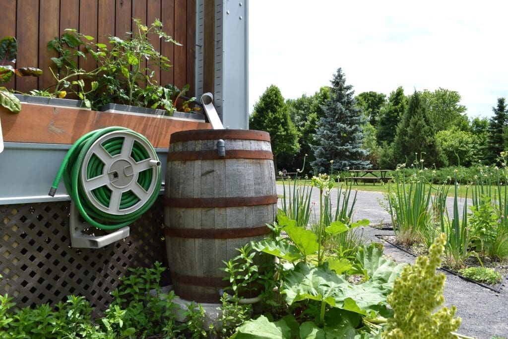 A wooden rainwater barrel in front of a house, surrounded by a garden and a forest