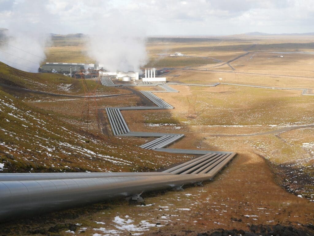 Geothermal power plant in a barren, open landscape, with steam rising from buildings and long insulated pipelines carrying hot water across the ground
