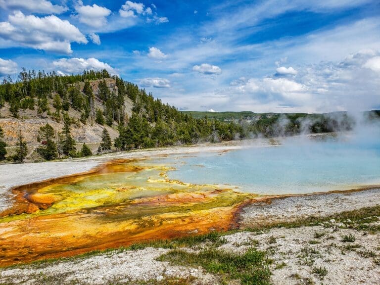 Geothermal hot spring with colorful mineral deposits in shades of yellow, orange, and green along the water’s edge, steaming under a blue sky with forested hills in the background