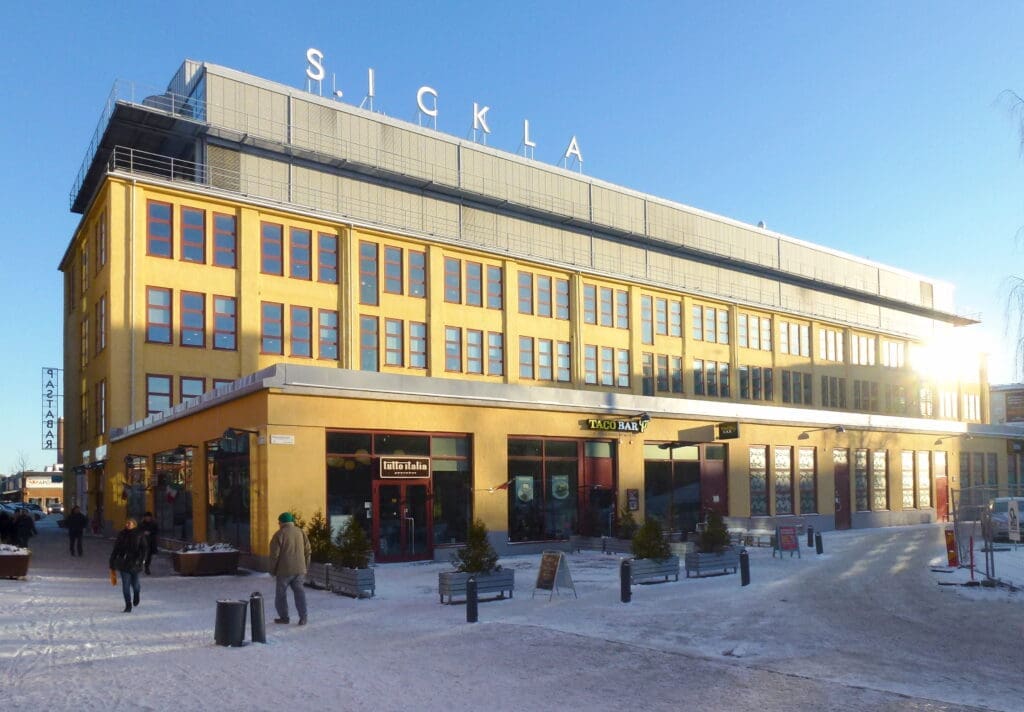 Large yellow industrial-style building with rows of windows, shops and cafés at street level, and pedestrians walking across a snowy plaza in bright winter sunlight. At the top of the building, large letters spell “SICKLA.”