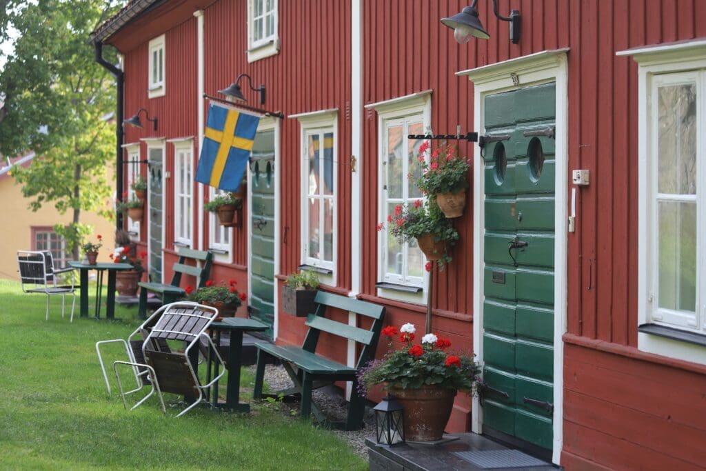 Row of traditional red wooden houses with green doors and white window frames, decorated with flower pots and benches; a Swedish flag hangs by one entrance, with garden chairs and greenery creating a calm residential setting.