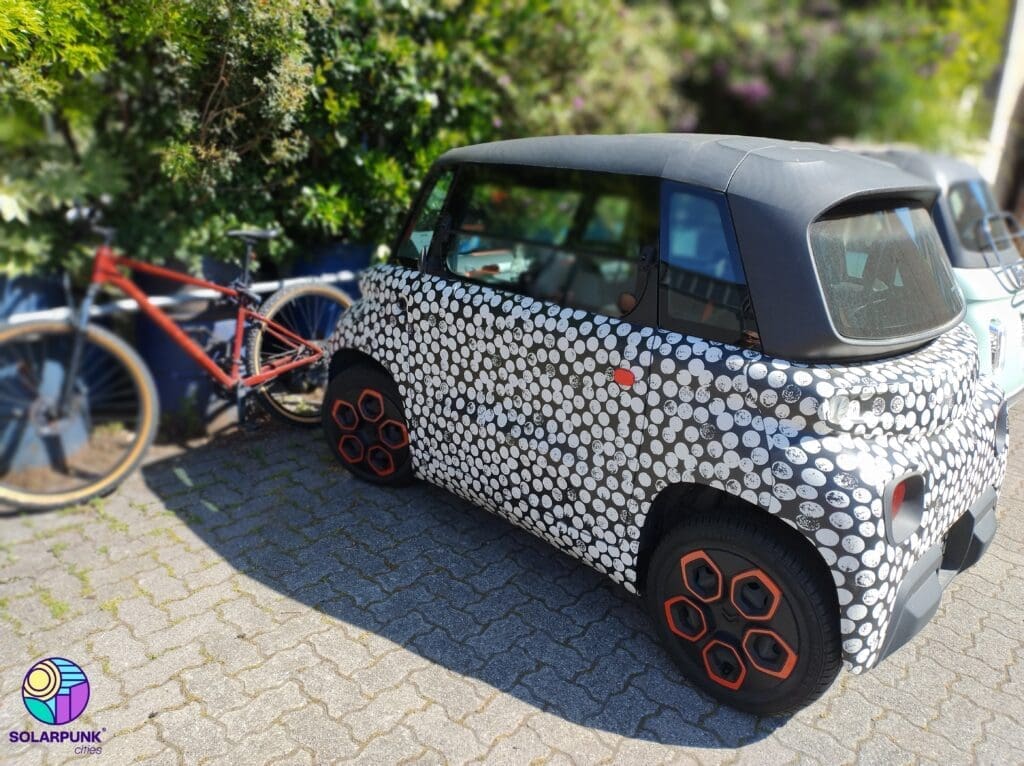 Compact electric car with a dot-patterned exterior parked on a residential street next to a bicycle, surrounded by greenery