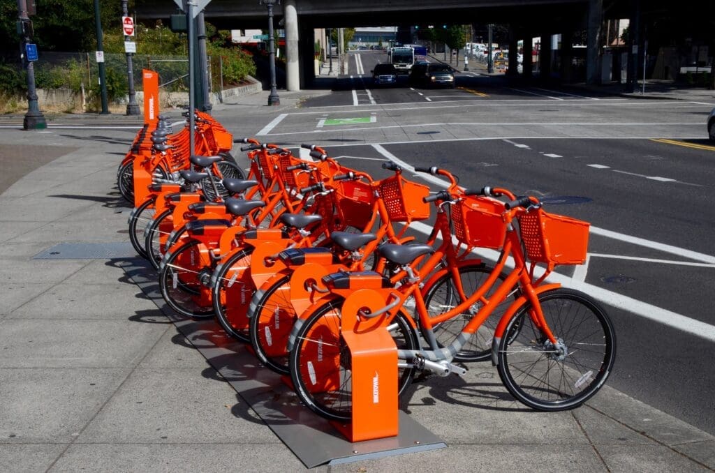 Bright orange bike-share bicycles are docked beside a protected bike lane on a city street, with an elevated roadway and light traffic in the background