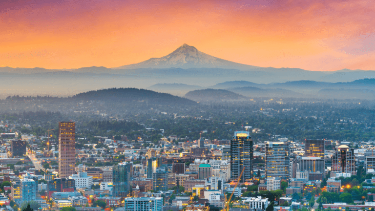 Portland, Oregon skyline at sunset, with downtown buildings in the foreground and snow-capped Mount Hood towering above layers of forested hills in the distance