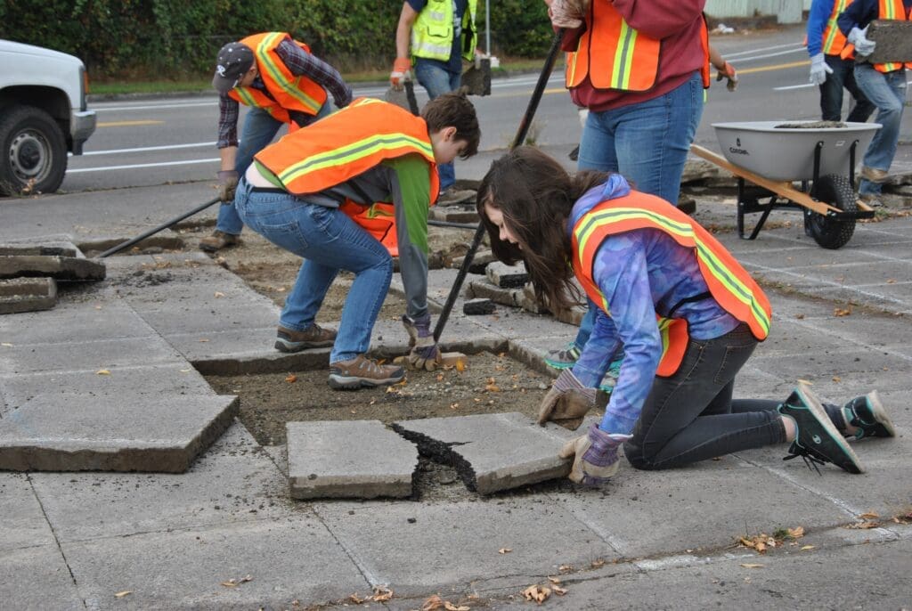 Volunteers wearing high-visibility vests remove sections of concrete pavement by hand using pry bars, shovels, and wheelbarrows, preparing the area for future greenery and improved stormwater management