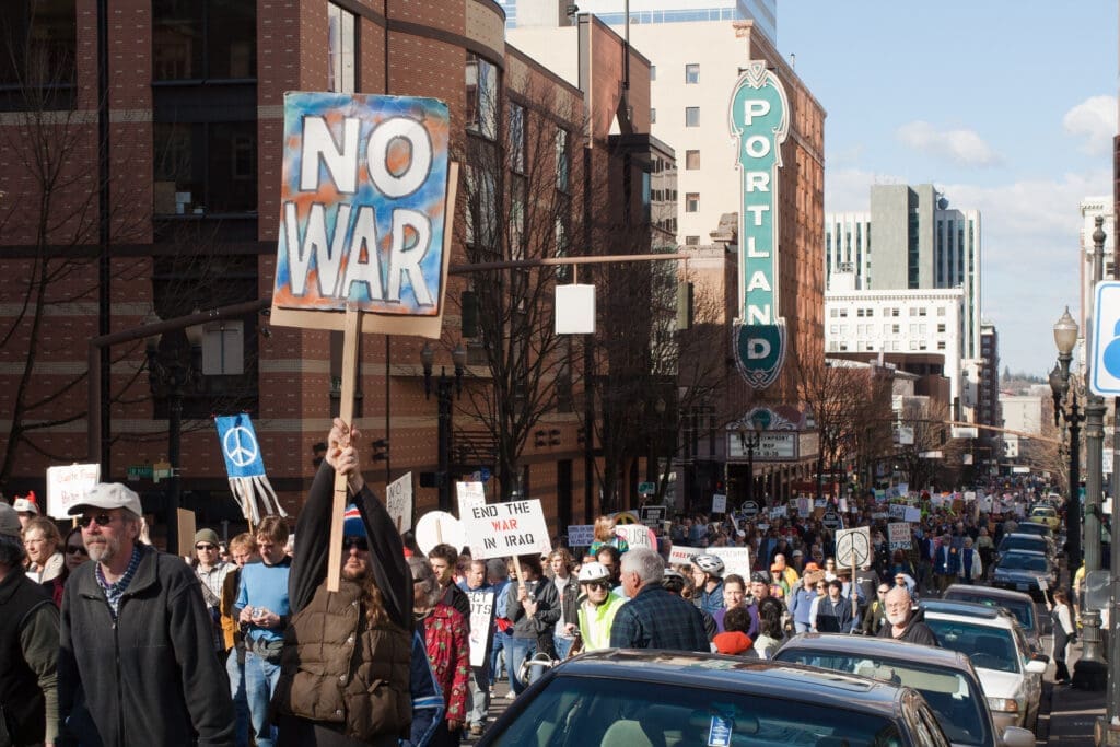 Anti-war demonstration in downtown Portland, with marchers carrying peace signs and protest banners. A large “NO WAR” sign is visible in the foreground beneath the iconic Portland theater sign.